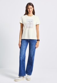 Femme debout portant un t-shirt blanc avec le texte "OWN IT", un jean bleu, des baskets blanches, et un bracelet noir au poignet droit.