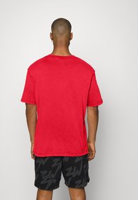 Camiseta de algodón roja de manga corta, con cuello redondo y un corte holgado, combinada con pantalones cortos negros que presentan un patrón de logo repetido.