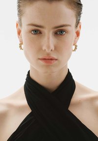 Top negro envolvente con textura suave, que presenta el rostro de una modelo, ojos azules y pendientes de aro dorados con acentos decorativos.