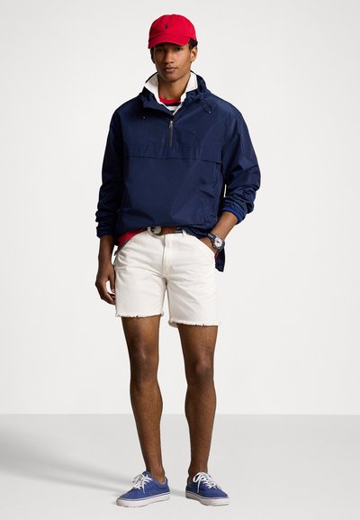 Veste coupe-vent marine avec fermeture éclair et capuche, associée à un short blanc effiloché et des baskets bleues. Une casquette rouge complète le look.
