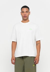 Biela tričko s krátkym rukávom a zeleným Adidas trefoil logom na ľavej hrudi, okrúhly výstrih, v kombinácii s olivovými nohavicami.
