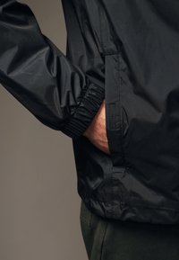 Ręka w kieszeni czarnej, lekkiej kurtki z elastycznymi mankietami, noszonej na ciemnozielonych spodniach, na jednolitym, neutralnym tle.