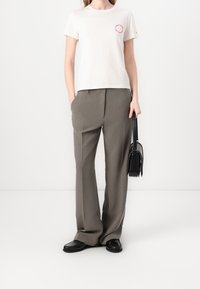 White cotton t-shirt with a round graphic logo on the chest, paired with high-waisted, wide-leg gray trousers and black shoes.