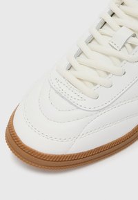 Άσπρο δερμάτινο αθλητικό παπούτσι με υφή στην επιφάνεια, με σόλα από καουτσούκ χρώματος καραμέλας, επίπεδ�α άσπρα κορδόνια και διακριτικές ραφές. Στρογγυλεμένο σχέδιο μύτης.