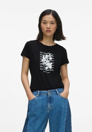 Młoda kobieta z krótkimi ciemnymi włosami nosi czarny T-shirt z nadrukiem i szerokie niebieskie dżinsy, ręce w kieszeniach, na jednolitym białym tle.