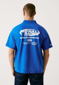 Ljusblå kortärmad pikétröja med en stor vit "RED BULL RACING"-logotyp och text på baksidan. Slät textur och avspänd passform.