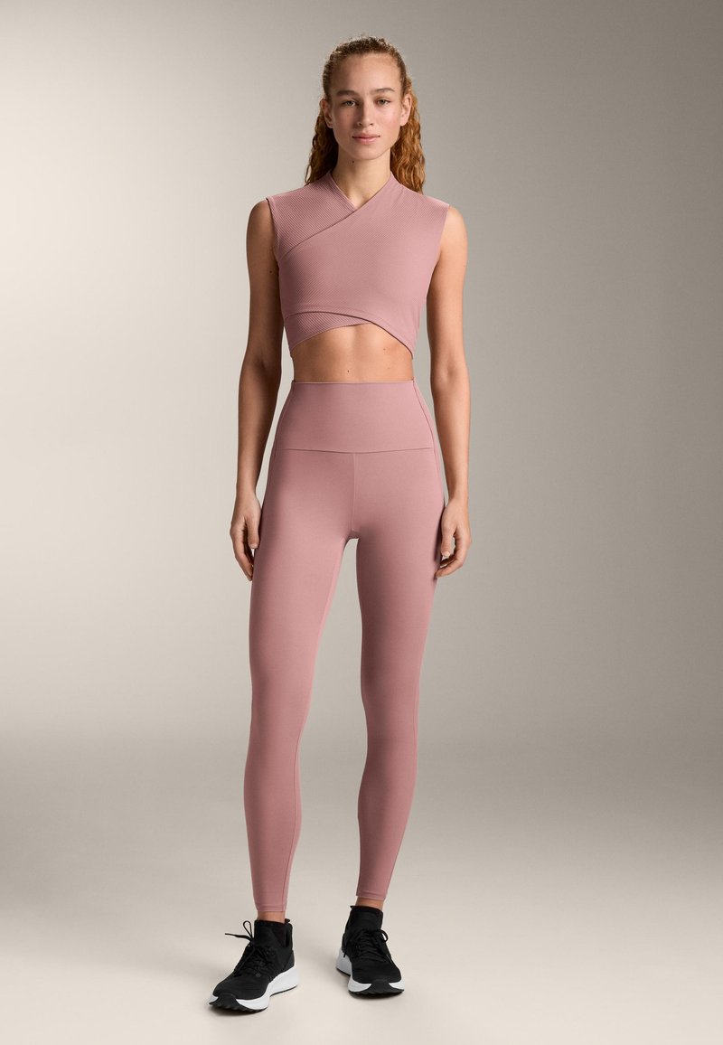 Set de haine active roz deschis format dintr-un top scurt, fără mâneci, cu design încrucișat și colanți cu talie înaltă, asortat cu pantofi sport negri.