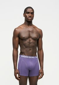 Μυώδης άνδρας στέκεται γυρνώντας προς τα εμπρός φορώντας μοβ μποξέρ Björn Borg μπροστά σε απλό φωτεινό φόντο.