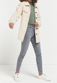 Beige fleece-takki nappilukolla, yhdistetty vihreään t-paitaan ja harmaisiin skinny-farkkuihin, täydentää valkoiset vetoketjulliset lenkkarit.