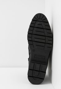 Fondo di scarpe da ginnastica nere con una suola in gomma testurizzata, scanalature profonde e un dettaglio di branding rettangolare. Design angolare con presa robusta.