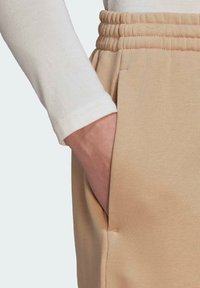 Beżowe dresy z bocznymi kieszeniami, z teksturowanym elastycznym paskiem w talii. Na jednym ramieniu widoczne mankiety białej koszuli z długim rękawem.