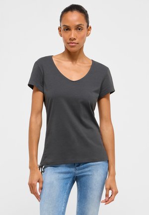 Kvinde iført en mørkegrå kortærmet V-hals t-shirt og lyseblå jeans, stående mod en ensfarvet lys baggrund.