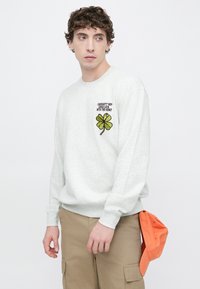 Hellgrauer Pullover mit rundem Halsausschnitt und langen Ärmeln, mit einem aufgestickten vierblättrigen Kleeblatt-Design und Text auf der linken Brust.