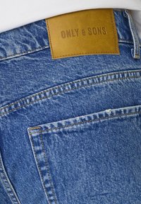Jeans de mezclilla azul claro con una textura suave, que cuentan con un parche de marca de cuero marrón y costuras en contraste en el bolsillo trasero y la cintura.