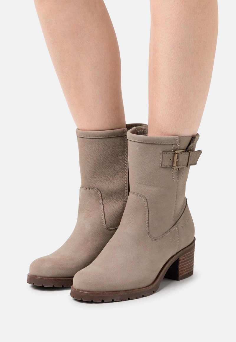 Bottes en cuir beige mi-mollet avec boucle latérale et talons carrés portées par une personne aux jambes nues sur un fond uni.