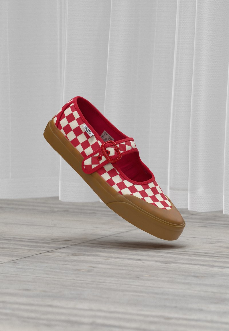 Chaussure slip-on à damier rouge et blanc avec une semelle en caoutchouc marron et une bride à boucle rouge, flottant au-dessus d'un sol en bois avec des rideaux blancs en arrière-plan.