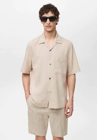 Lichtbeige korthangshirt met een borstzak en zwarte knopen, in combinatie met beige gestreepte shorts. Model draagt zwarte zonnebril.