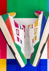 Paire de baskets blanches avec des panneaux en daim multicolores verts, jaunes, rouges, bleus et roses, positionnées semelle contre semelle sur un fond coloré.