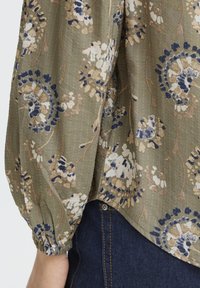 Oliiviroheline pluus kreemja, sinise ja pruuni lillemustriga. Omab elastseid mansette ja tekstuurset kangast. Kaardus alläär.