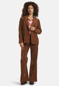 Brown corduroy suit featuring a tailored blazer and flared pants, paired with a pink patterned shirt. Textured fabric and structured design.