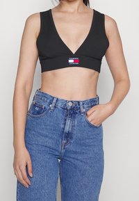 Svart ribbad crop-top med djup V-ringning och en Tommy Jeans-logotyp i mitten. Kombinerad med högmidjade blå denimjeans.