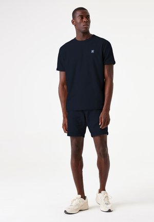 Navyblaues Baumwoll-T-Shirt mit kurzen Ärmeln und kleinem Logo auf der Brust, kombiniert mit navyblauen Shorts und weißen Turnschuhen.