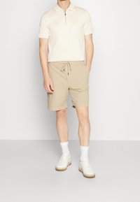 Písková polokošile s zipem u límce v kombinaci s pískovými šortkami s elastickým pasem. Model má na sobě bílé tenisky a bílé krátké ponožky.