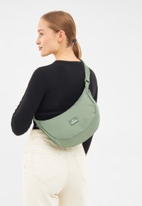 Borsa a spalla in tessuto verde con forma morbida e curvilinea, dotata di tracolla regolabile, con un'etichetta sul davanti. Indossata sopra una maglietta nera e pantaloni bianchi.