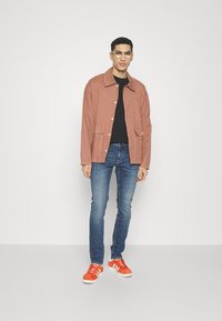 Chaqueta casual de color coral claro con dos bolsillos frontales, combinada con una camiseta negra, jeans azules y zapatillas naranjas con detalles blancos.