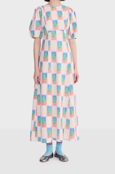 Femme portant une longue robe à manches bouffantes avec un motif géométrique coloré, associée à des chaussettes bleu clair et des ballerines métalliques.