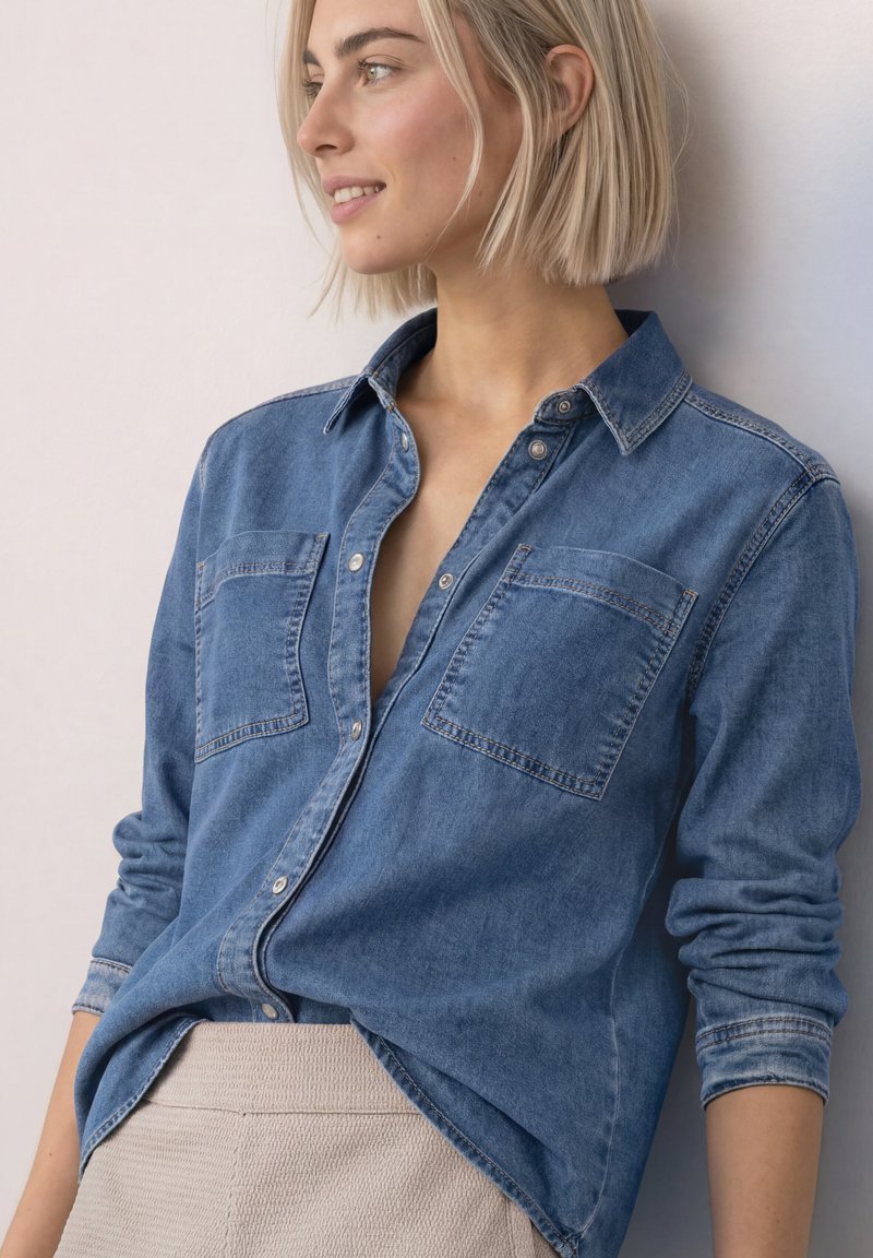 Femme blonde aux cheveux courts portant une chemise en jean bleue et un pantalon beige, appuyée contre un mur clair, regardant sur le côté en souriant.