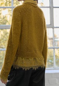 Senfgelber strukturierter Cardigan mit kurzem Schnitt, getragen über einer Bluse mit Spitzenbesatz. Schwarze Lederhose komplettiert das Outfit.