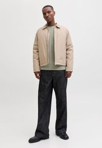 Mann trägt beigefarbene Reißverschlussjacke, hellgrünes Hemd, lockere dunkle Jeans und schwarze Schuhe, steht vor hellem einfarbigem Hintergrund.