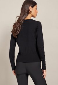 Svart tröja med långa ärmar, ribbade muddar och figursydd midja. Har guldknappdetaljer vid mudden. Mjuk och slät textur.