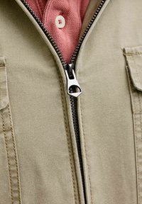 Beige Jacke, halb über ein rotes Hemd mit sichtbarem weißen Knopf gezippt, zeigt Stoffstrukturen und Details des Metallreißverschlusses.