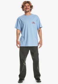 Camiseta de algodón azul claro con un pequeño gráfico rosa y blanco en el pecho, combinada con pantalones anchos oscuros y estampados y zapatos negros.