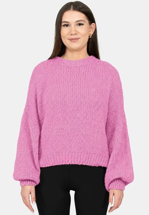 Junge Frau mit langen braunen Haaren, die einen pinken grob gestrickten Pullover und schwarze Hosen trägt, steht vor einem schlichten hellen Hintergrund.