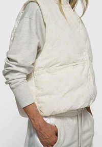 Weiße gesteppte Weste mit dezentem Logomuster, ausgestattet mit einer Reißverschlusstasche, Druckknöpfen und einem hohen Kragen. Trägt man über einem hellgrauen Sweatshirt.