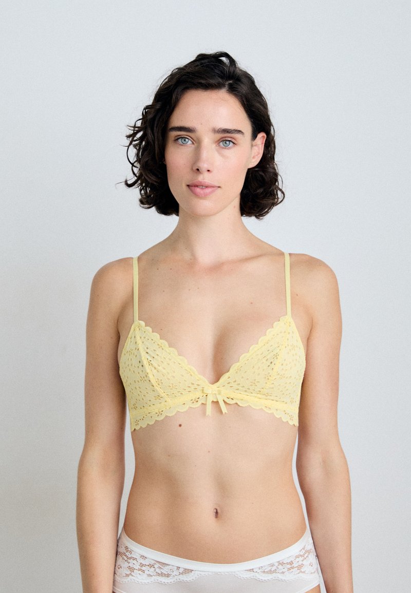 Bralette giallo in pizzo con bordo ondulato, caratterizzato da un dettaglio a fiocco al centro e spalline sottili regolabili.