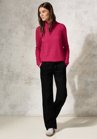 Fuchsia gestrickter Pullover mit hohem Kragen, kombiniert mit lässigen schwarzen Hosen und grauen Sneakers mit weißen Akzenten vor einem strukturierten Hintergrund.