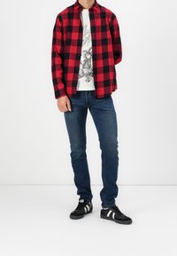 Hombre vestido con una camisa de cuadros rojo y negro sobre una camiseta gráfica blanca, jeans azules y zapatillas negras con rayas blancas, de pie contra un fondo blanco.