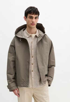 Młody mężczyzna w taupe kurtce z kapturem, na beżowej, paskowanej koszuli zapinanej na guziki oraz jasnych spodniach, stoi na białym, jednolitym tle.