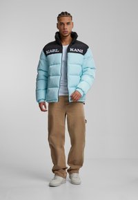 Lyseblå jakke med fyld, sort krave og teksten "KARL KANI". Sammen med beige bukser og hvide sneakers. Glat materiale, quiltet design.