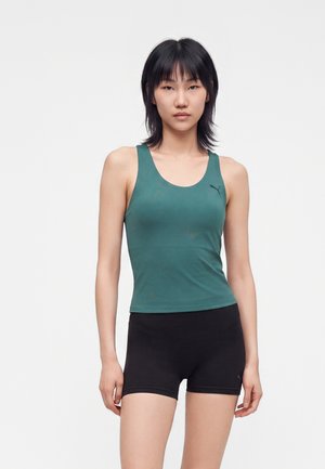 Jeune femme aux cheveux noirs porte un débardeur vert sans manches et un short noir moulant, debout devant un fond clair uni.