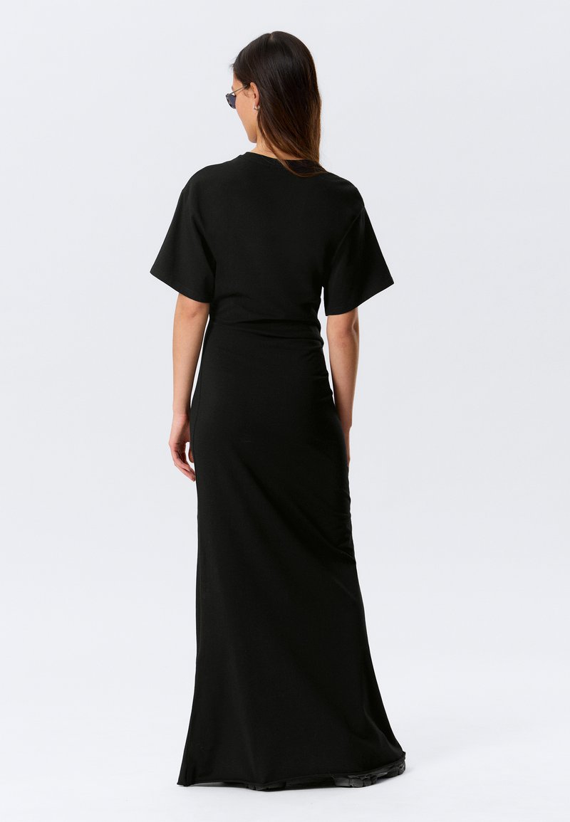 Moteris ilgais plaukais, dėvinti juodus akinukus nuo saulės ir aptemptą juodą maksi su trumpomis rankovėmis suknelę, stovinti nugara į baltą foną.