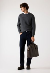 Maglione grigio scuro texture, jeans blu scuro, borsa in pelle marrone con hardware dorato, scarpe scure, in piedi in modo informale contro uno sfondo chiaro.