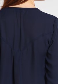 Marineblå bluse med en glat tekstur, der har et diskret bærestykke-design og syninger på ryggen. Lange ærmer og en afslappet pasform.