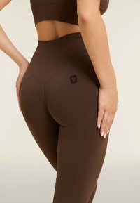 Leggings marroni a vita alta con una finitura testurizzata e un dettaglio del logo sulla coscia superiore, caratterizzati da un design attillato e cuciture accentuate.