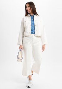 Weiße Jeansjacke über einem blauen Hemd mit Knöpfen, kombiniert mit weißen weiten Hosen und silbernen Sneakern, während sie eine Canvas-Tasche mit blauen Akzenten hält.