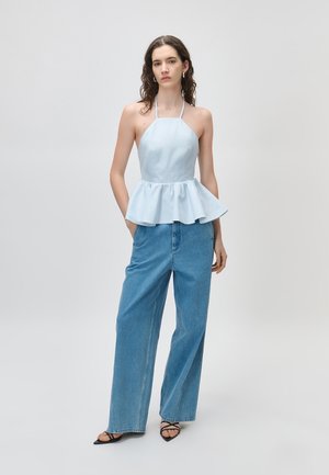 Donna con capelli ricci che indossa un top peplum azzurro chiaro con collo ad halter, jeans blu a gamba larga e tacchi neri con cinturini su uno sfondo semplice.